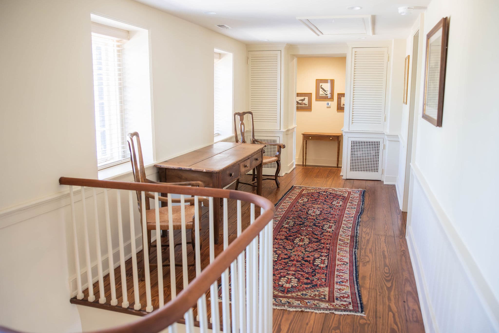 A well-lit hallway features a wooden table, chairs, and a decorative rug.