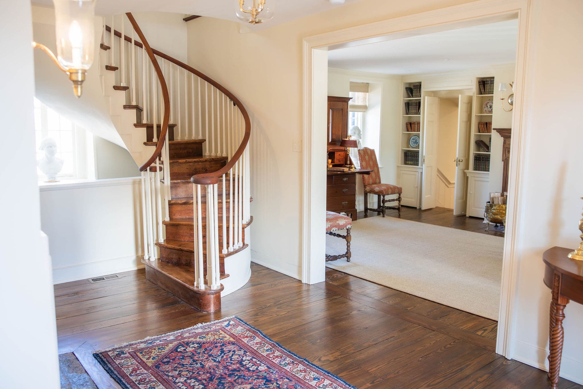 A warmly lit entrance hall features a curved wooden staircase, hardwood floors, and an adjoining room with furniture.