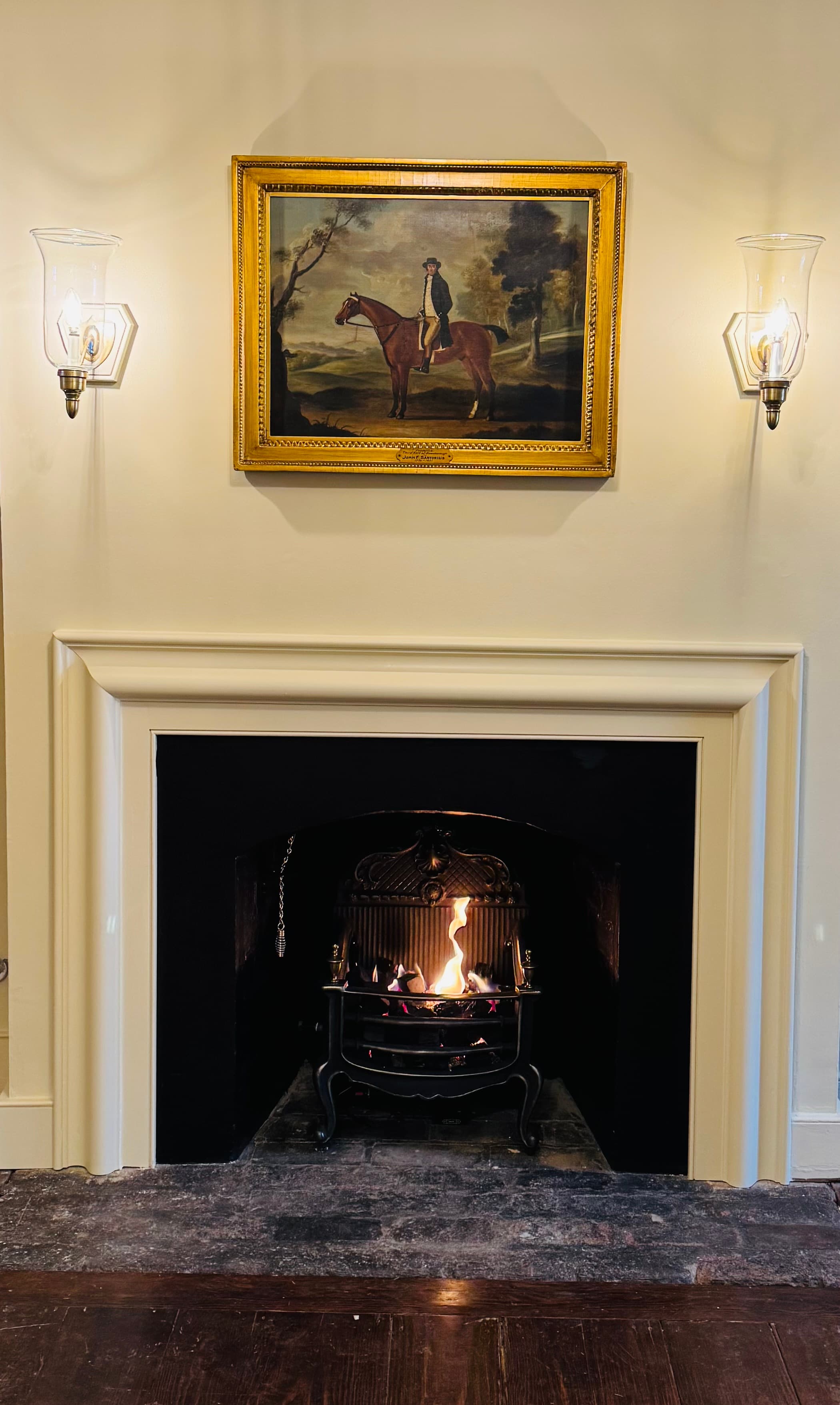 A classic fireplace with a lit fire, framed by wall sconces and a horse painting above.